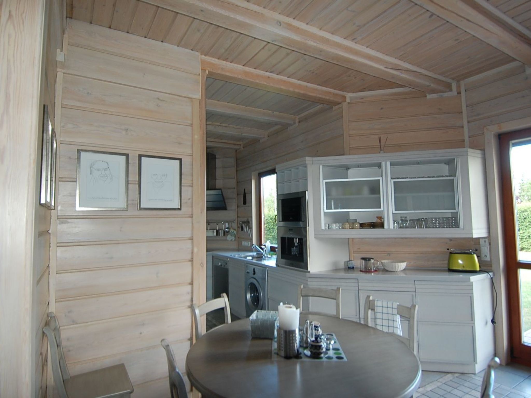 A wooden cabin interior with a dining table, chairs, a kitchen area with appliances, and framed artwork on the wall.