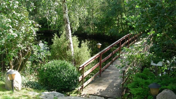 Un puente de madera con barandilla de metal, rodeado de abundante vegetación y un sendero de piedra.