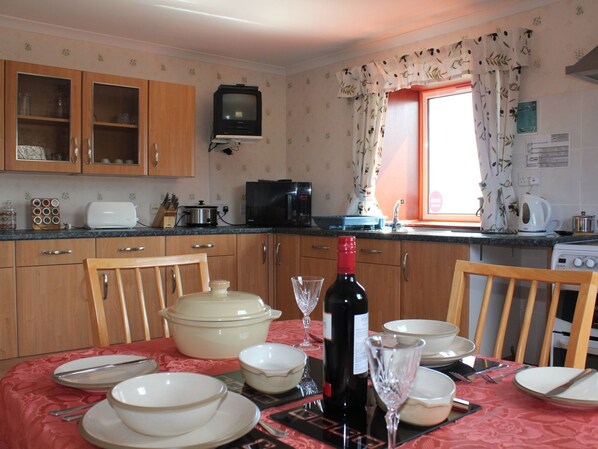 A kitchen with wooden cabinets, a microwave, a toaster, and a dining table set with plates, bowls, and a wine bottle. - Rustikales Ferienhaus Shiantview by Interhome (Orinsay)
