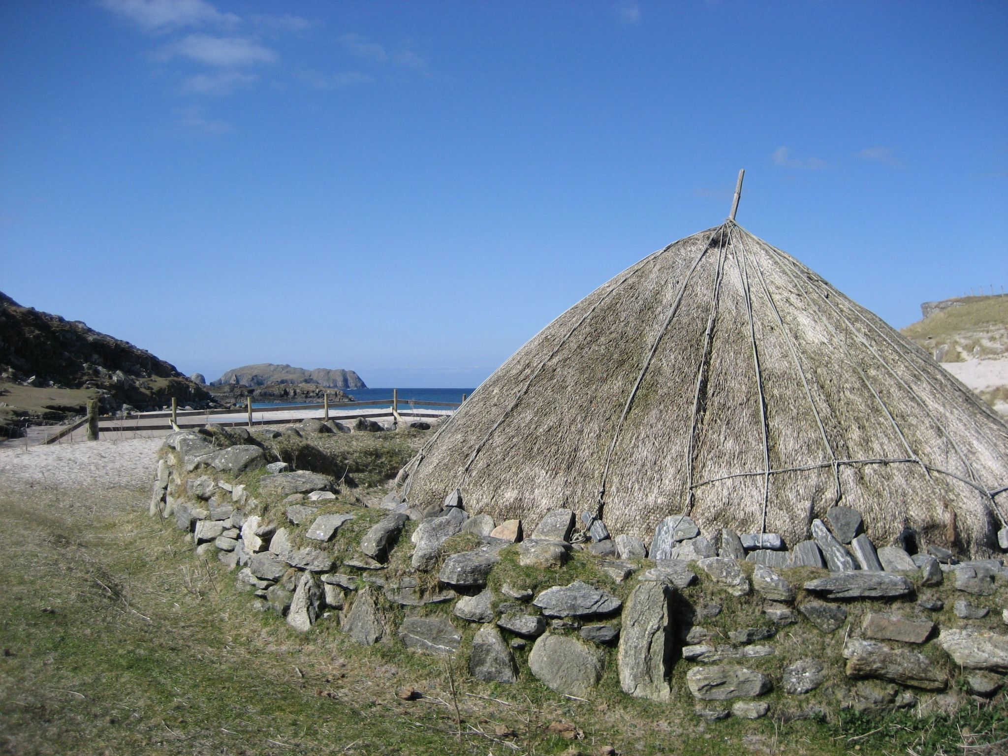 A traditional thatched-roof hut surrounded by a stone wall, set against a coastal backdrop with clear skies.