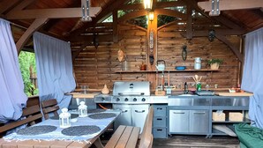 Une cuisine extérieure rustique, avec des tables et des bancs en bois, un barbecue et divers ustensiles de cuisine.