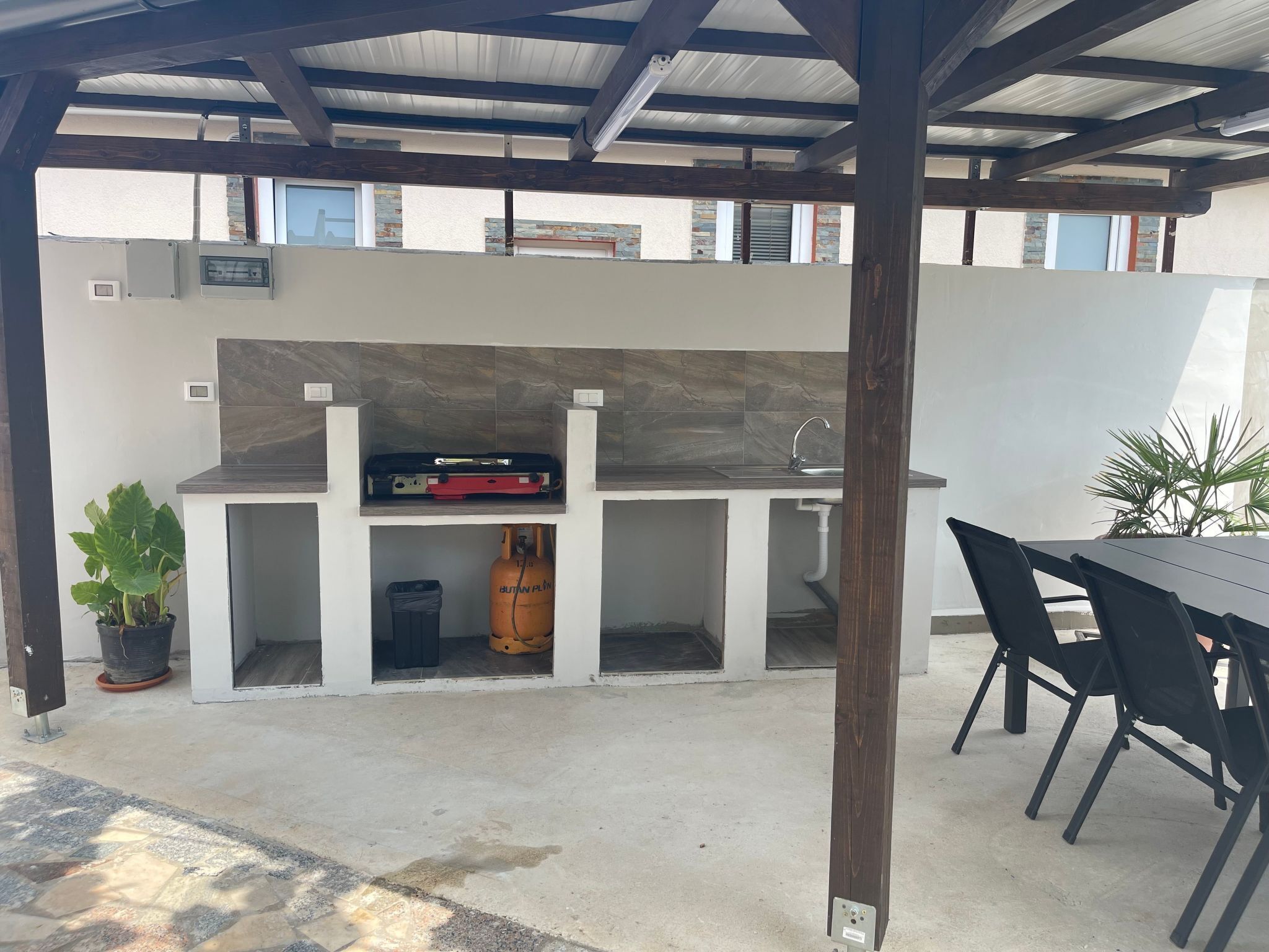 An outdoor kitchen area with a built-in grill, a sink, and a gas cylinder. There is a table and chairs to the right, and a potted plant to the left.