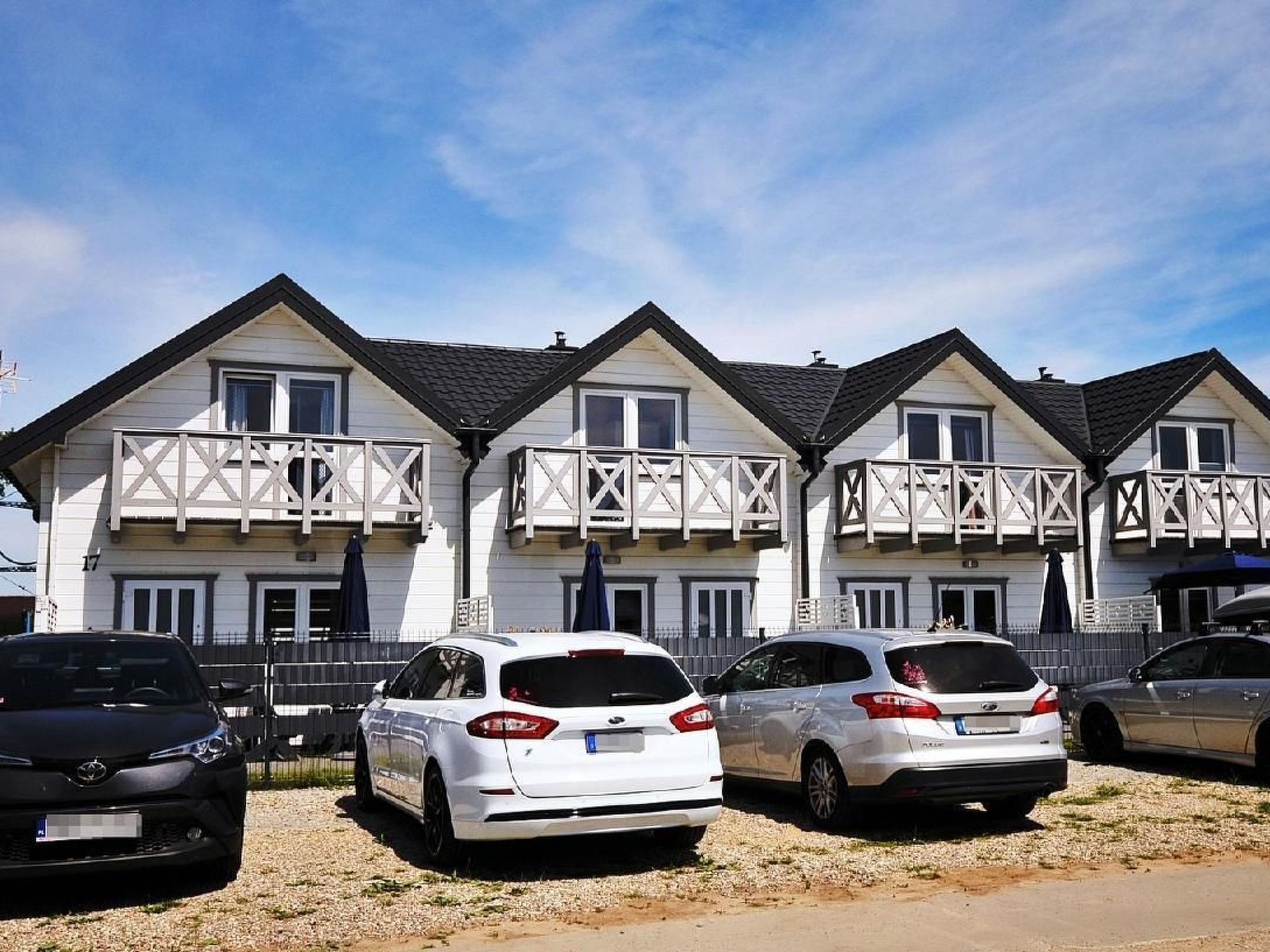 Une rangée de maisons blanches dotées de balcons, des voitures garées devant, et un ciel dégagé.