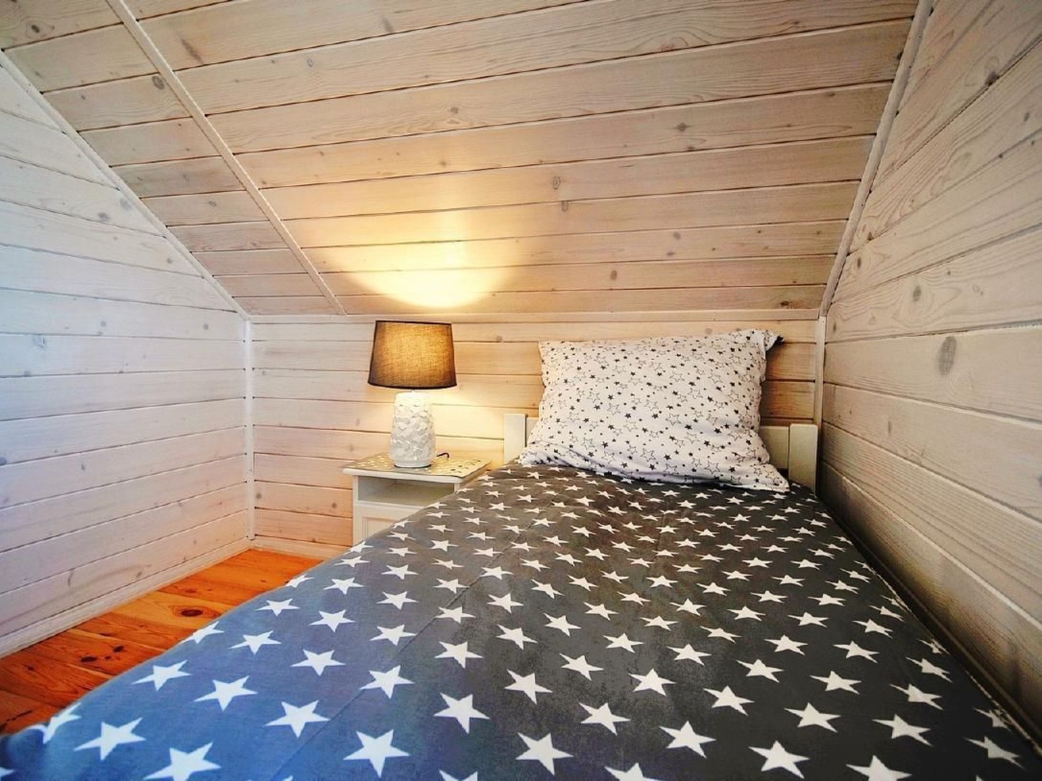 Een kleine, knusse kamer met houten wanden en een schuine kap. Er staat een bed met een deken met stermotief, een bedlampje en een kussen met sterrenprint.