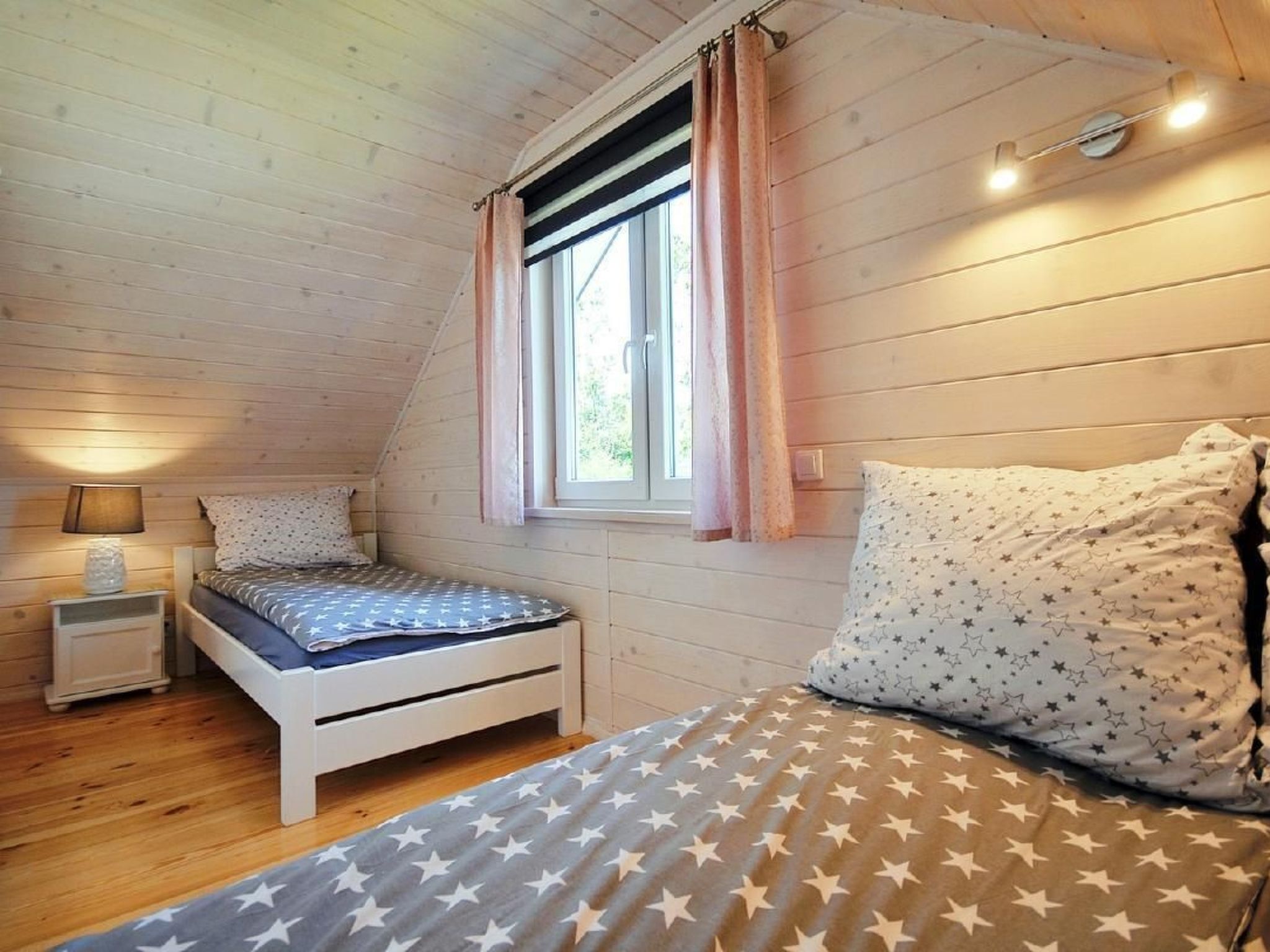 Une chambre mansardée chaleureuse, avec un plafond en pente, un lit simple recouvert d’une courtepointe à motifs d’étoiles, une lampe de chevet et une fenêtre avec des rideaux.