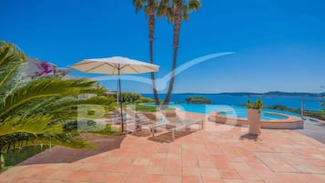 A poolside area with lounge chairs and an umbrella, surrounded by palm trees and a view of the sea.