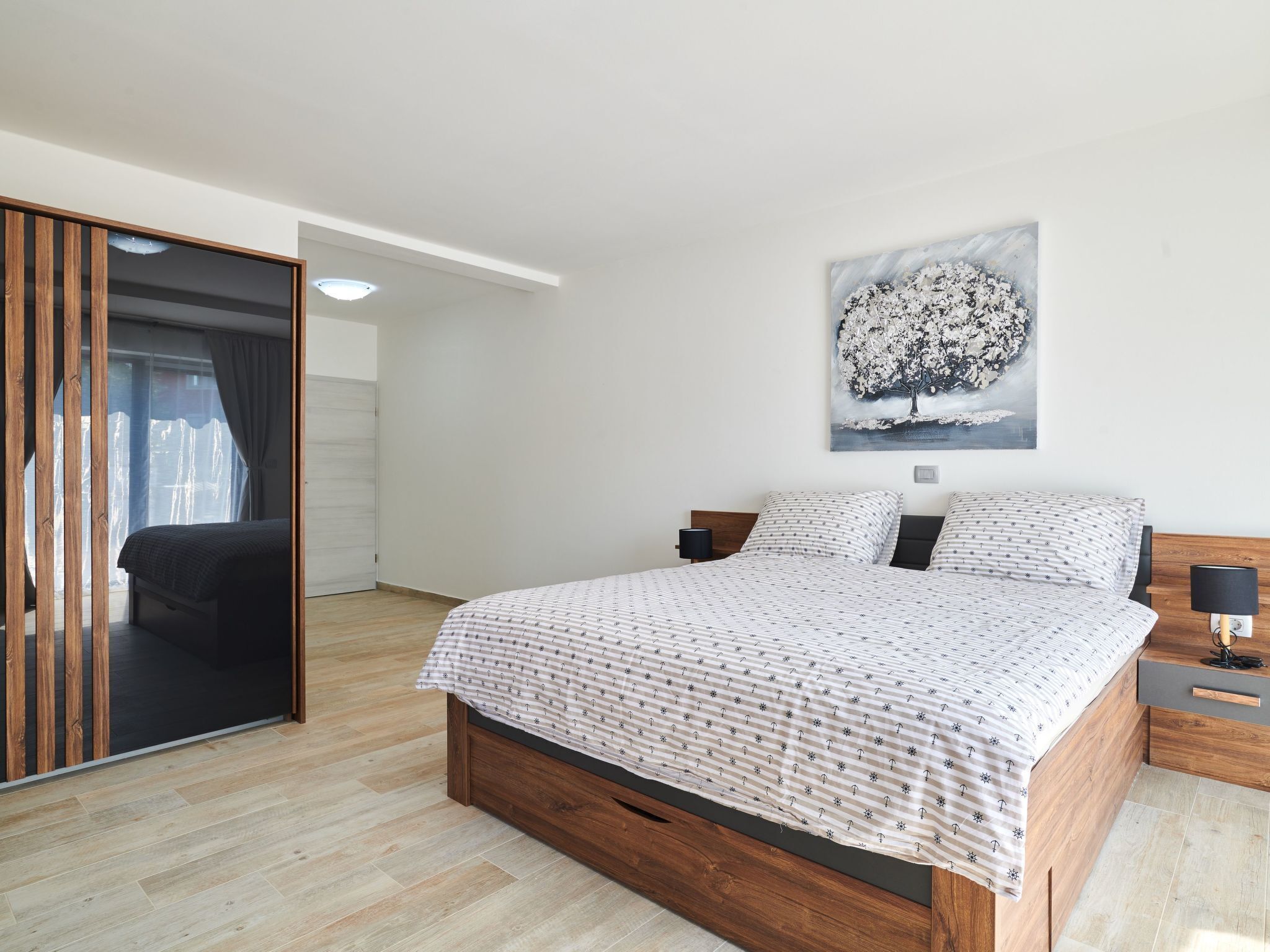 A modern bedroom with a wooden bed, a large window, and a painting of a tree.