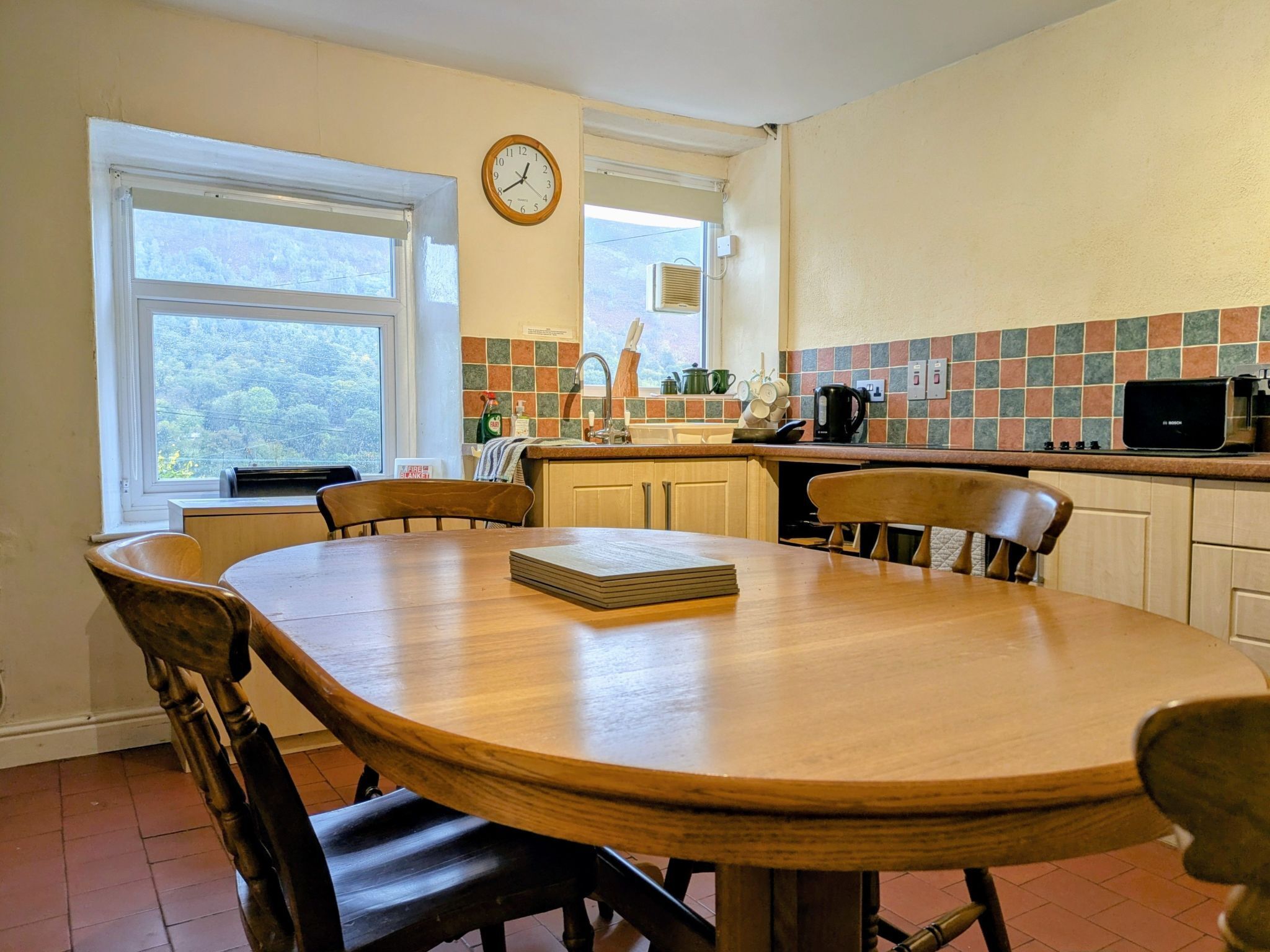 Une cuisine avec une table en bois, des chaises, une horloge et une fenêtre donnant sur des arbres.