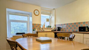A kitchen with a wooden table, chairs, a clock, and a window with a view of trees. - Velvet Cottage by Interhome (Pentrefelin)