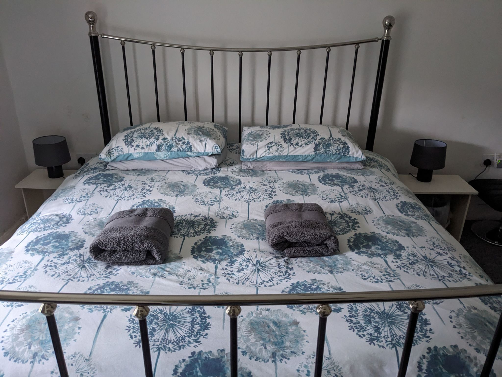 A single bed with a metal frame, two pillows with a floral pattern, two bedside lamps, and two folded towels on the bed.