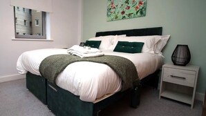 Ein ordentlich bezogenes Bett mit weißer und grüner Bettwäsche, ein Nachttisch mit Lampe und ein Fenster mit Jalousien.
