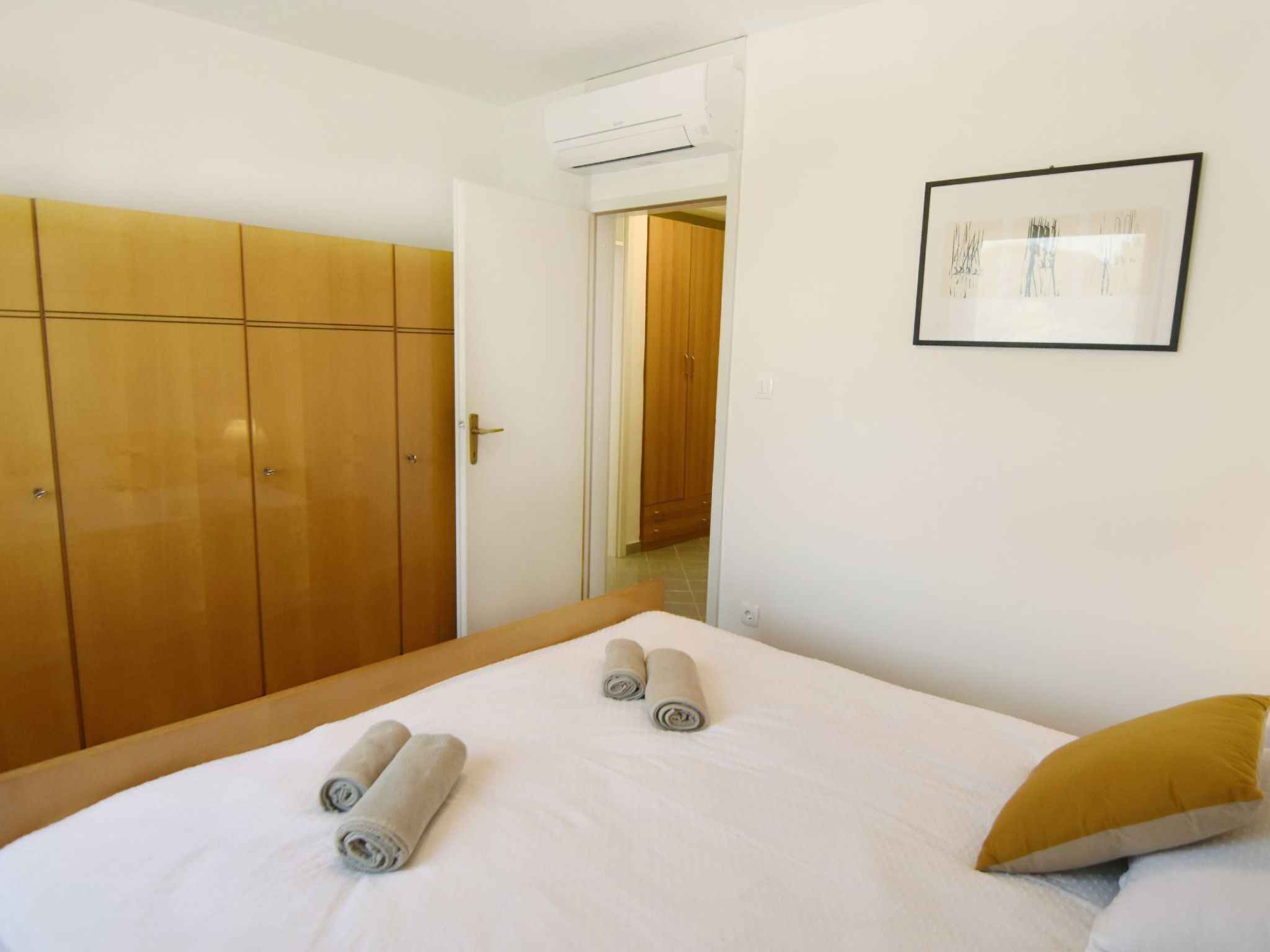 A hotel room with a bed, two rolled towels, a yellow pillow, a wardrobe, and a framed picture on the wall.