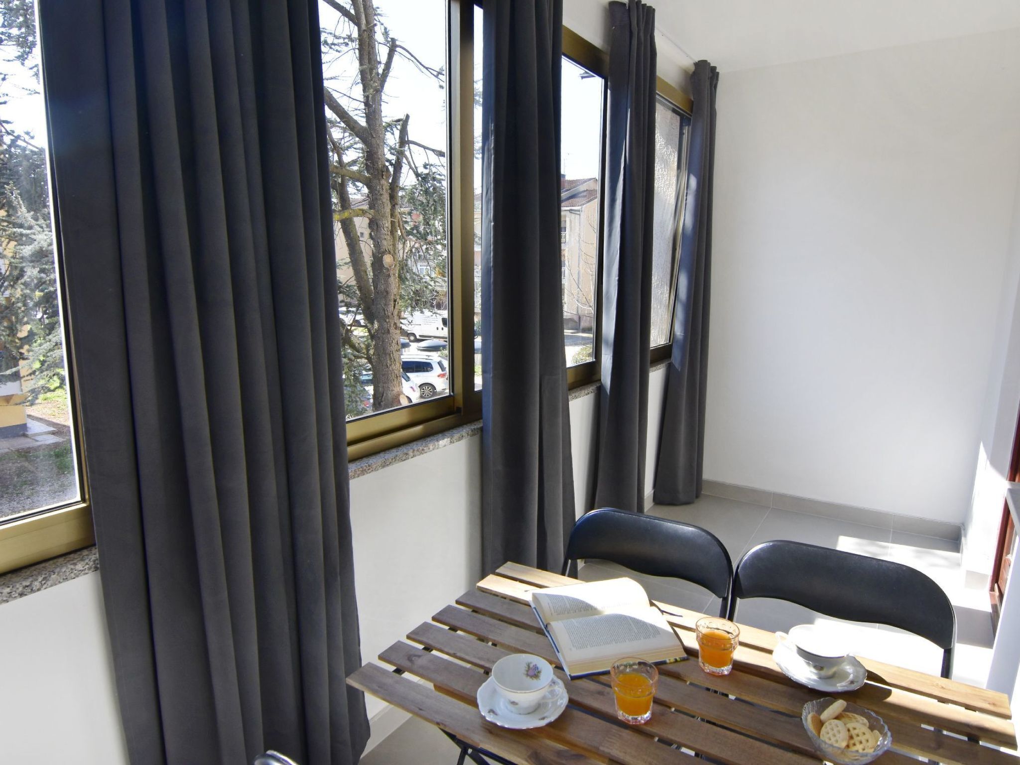 Una habitación con una mesa de madera lista para el desayuno, una ventana con cortinas y vistas a árboles y coches aparcados afuera.