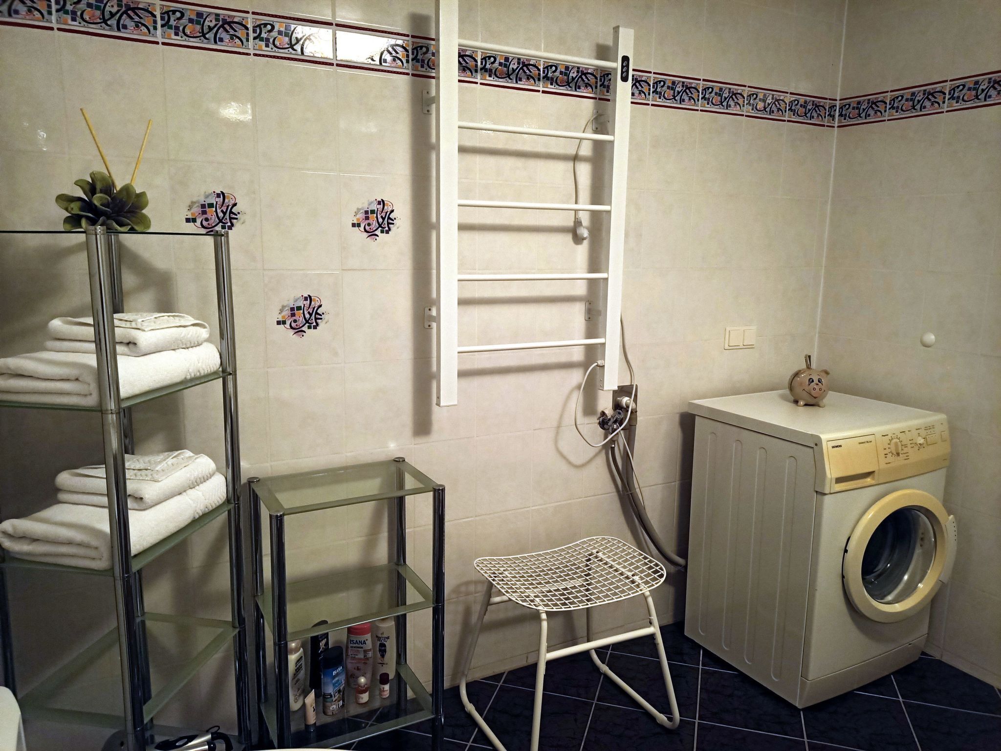 A bathroom with a washing machine, a metal shelf with folded towels, a small stool, and a wall-mounted rack.