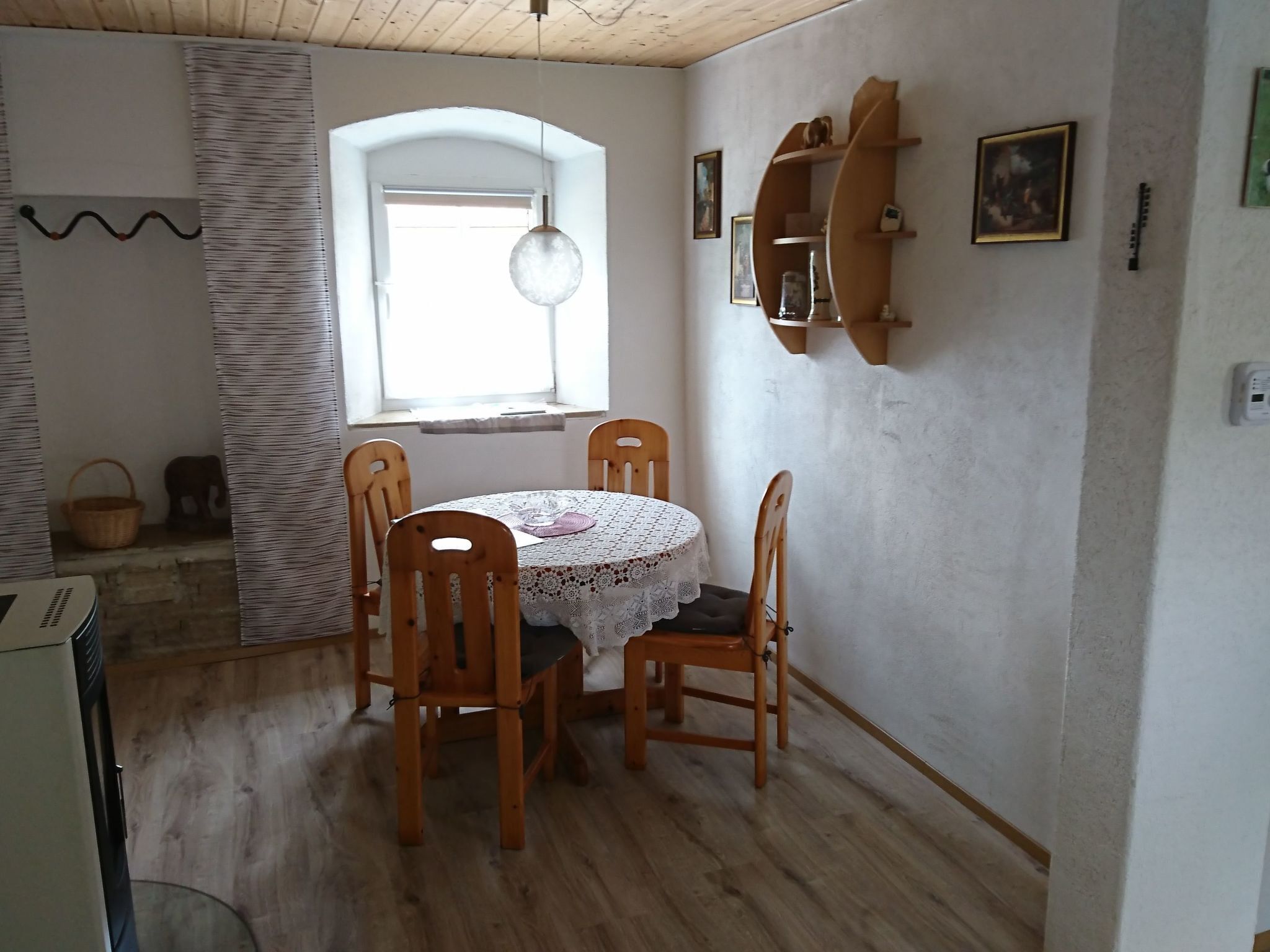 Ruokailutilassa on pyöreä pöytä ja neljä puista tuolia, ikkunassa roikkuu valaisin ja seinälle on kiinnitetty hylly, jossa on valokuvia.