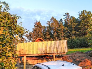 Une voiture blanche est garée devant une structure en bois, dont la base est en pierre, et l’ensemble est entouré d’arbres et de verdure.