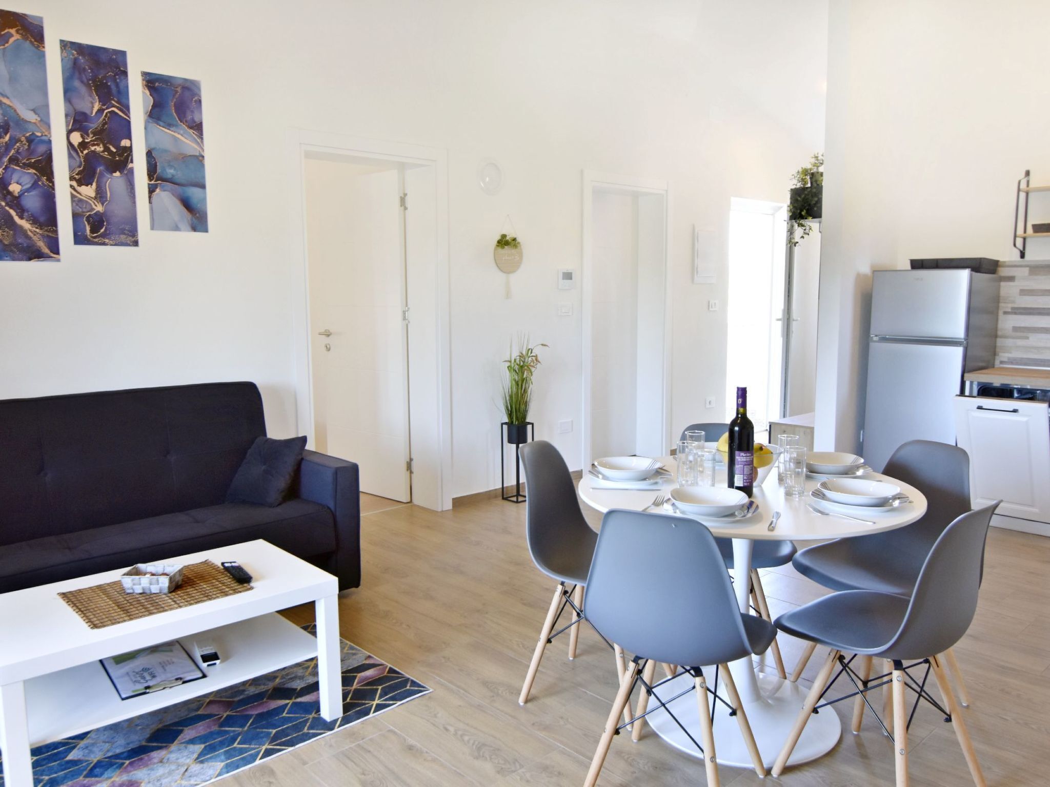 A modern living room with a dark sofa, a white coffee table, and a dining area with a round table and four chairs. There is a kitchen area with a refrigerator and a sink.