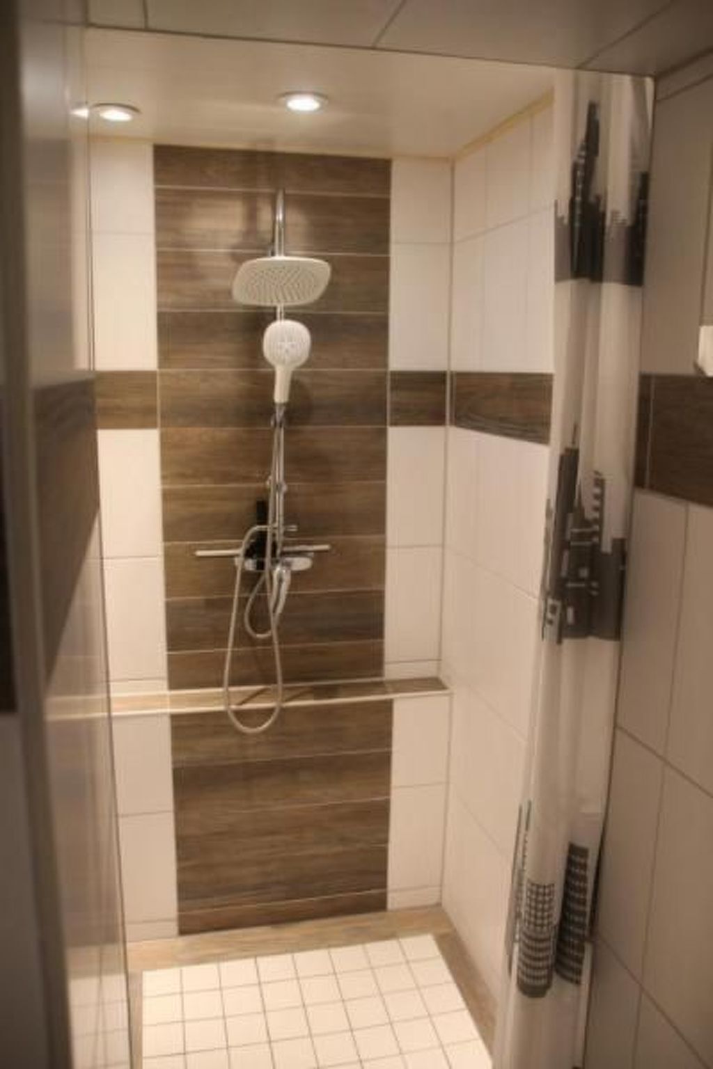 Eine Dusche mit Handbrause und am Wand befestigtem Wasserhahn, Fliesenwände mit Holzoptik und einer Glasduschwand.