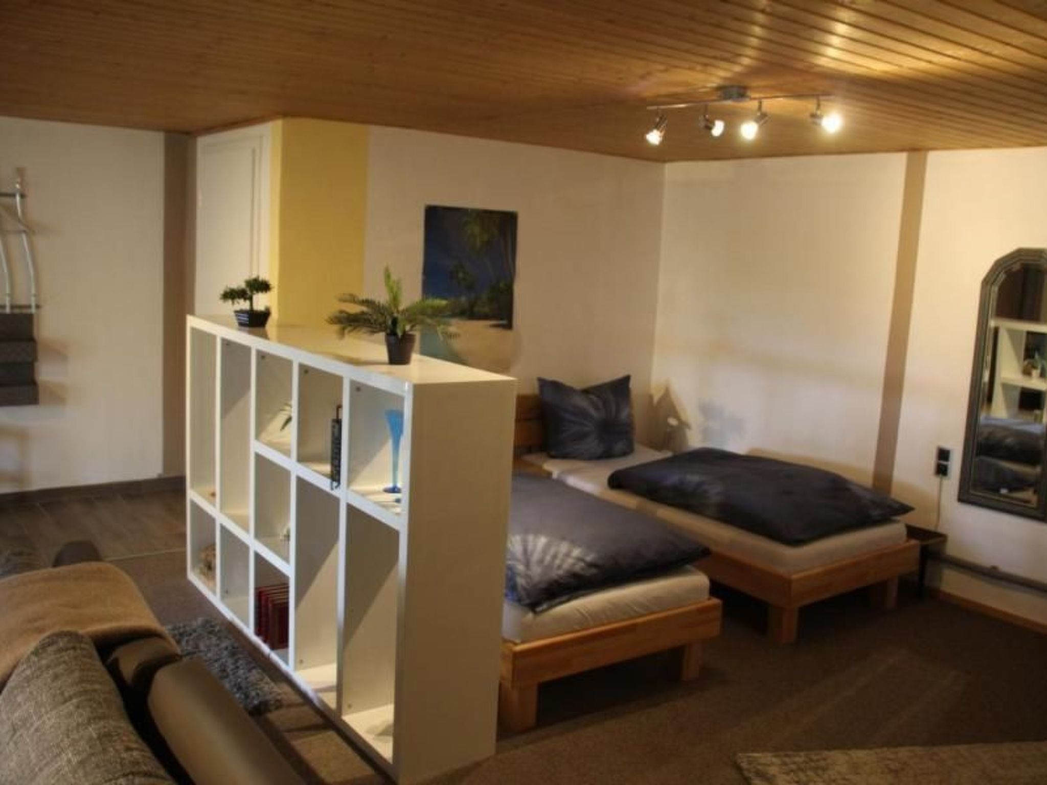 A compact room with a bed, a small seating area, and a white shelving unit with plants.