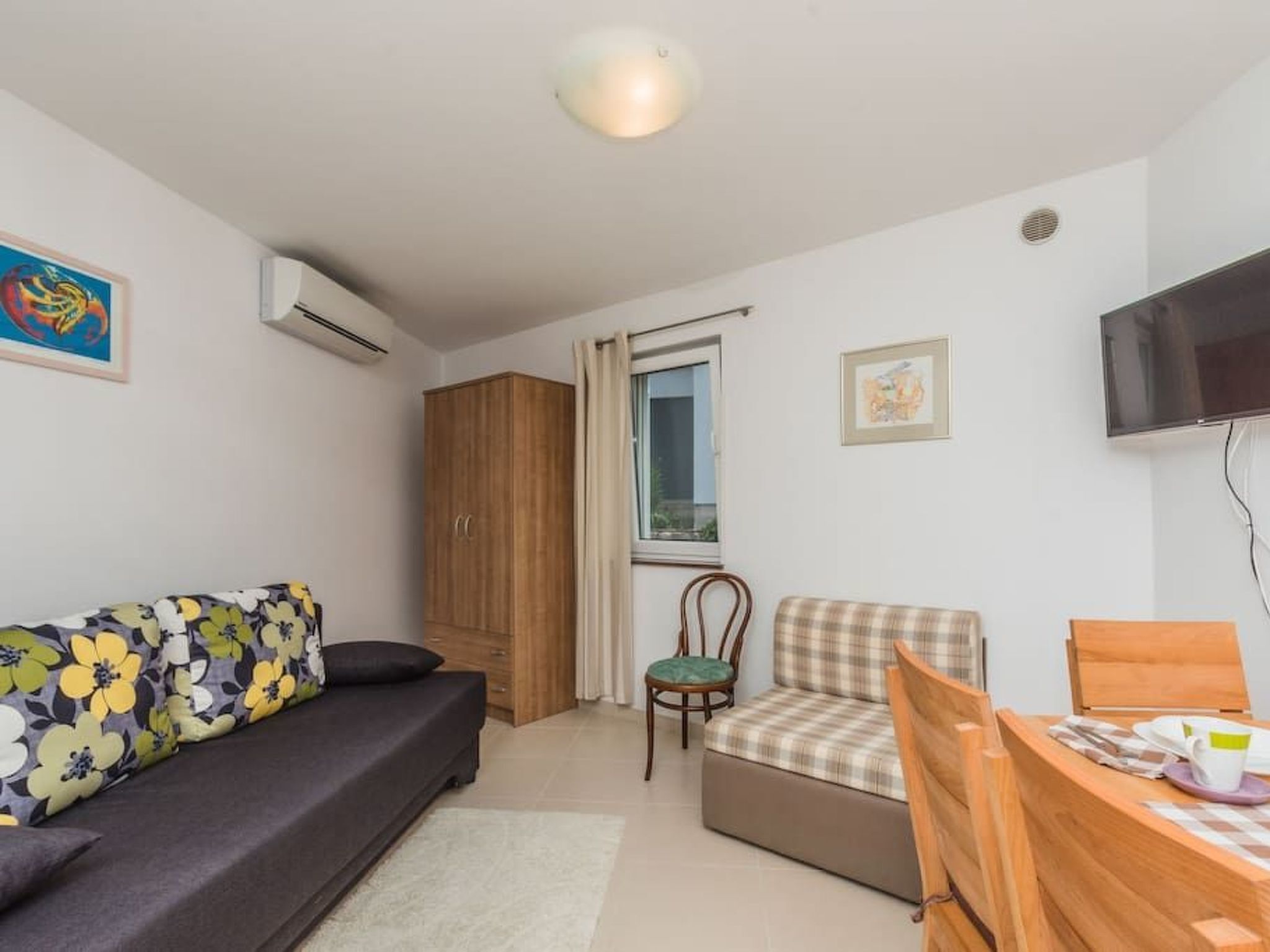 A compact living space with a sofa, a chair, a small table, a wooden wardrobe, a window with curtains, and a mounted television.