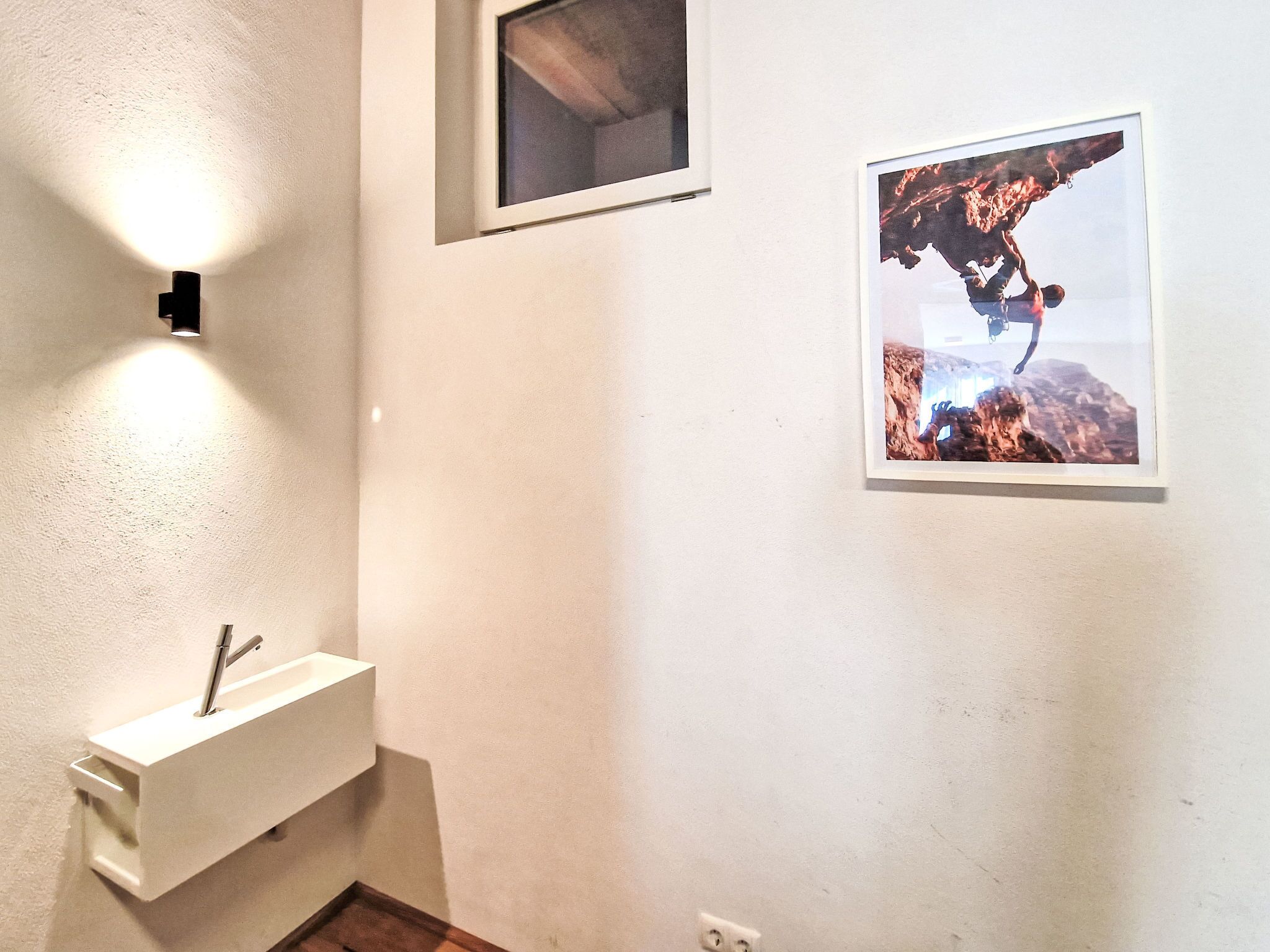 Une salle de bain avec un lavabo blanc, un luminaire fixé au mur et un tableau représentant une personne en train d’escalader un rocher.
