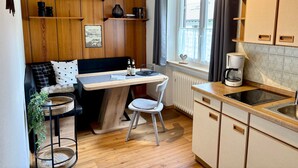 A compact kitchen with wooden cabinets, a dining area with a bench and chairs, a small window with curtains, and a radiator. - Haus Rosenwirth by Interhome (Mittenwald)