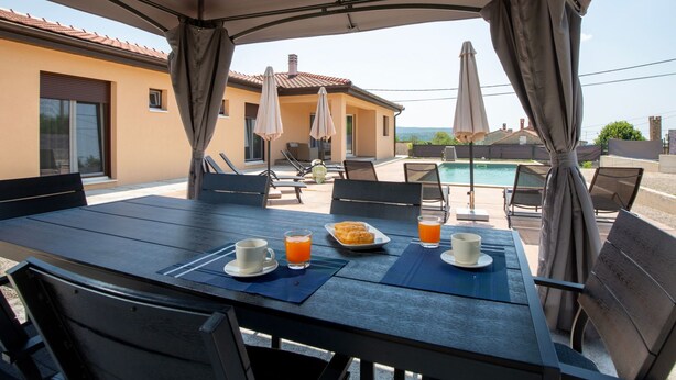 Een eethoek buiten met uitzicht op het zwembad, overdekt door een groot zonnescherm, met lounge fauteuils en parasols.