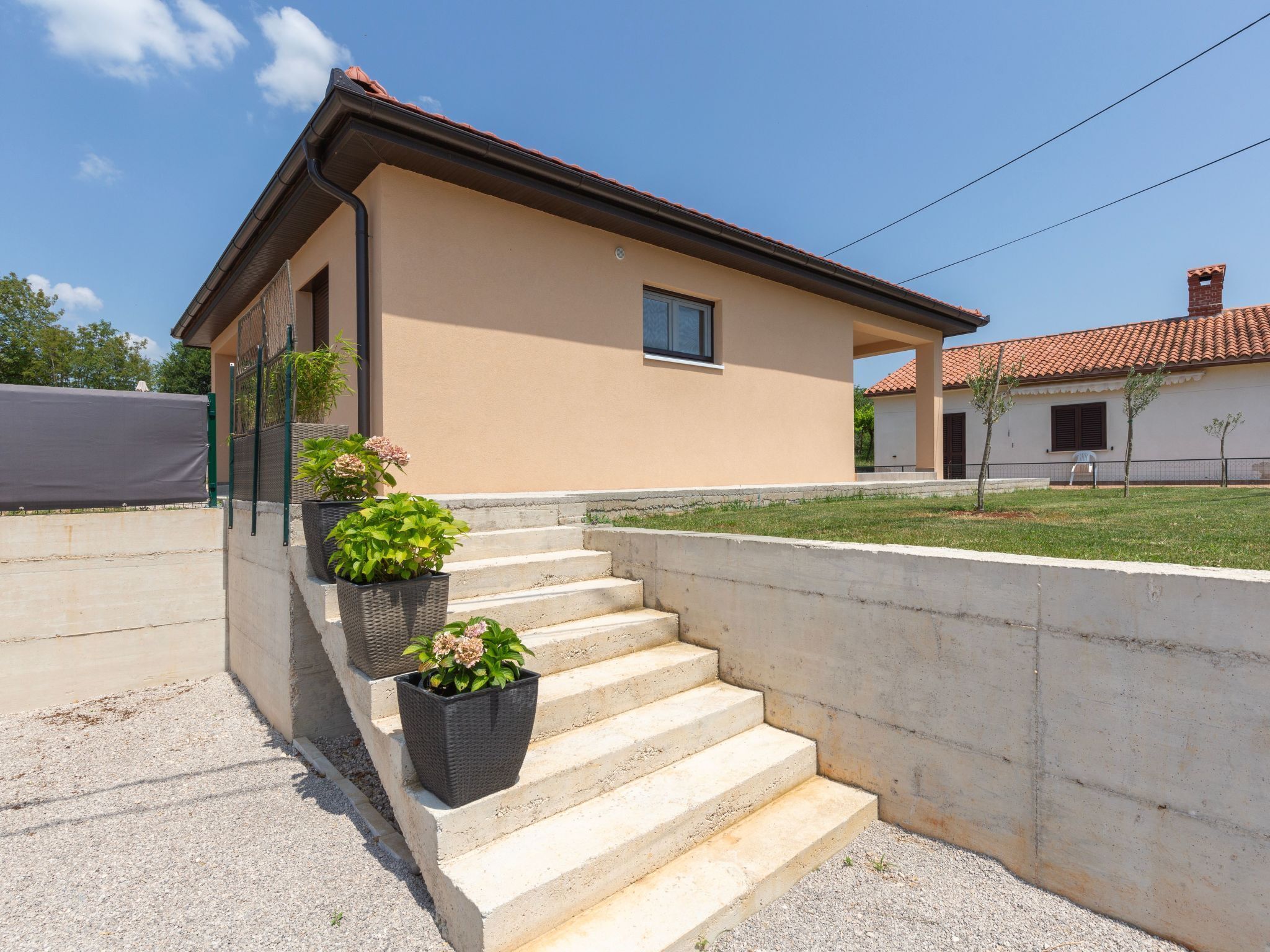 Una casa de dos pisos con fachada color beige, techo de tejas rojas y una escalera de concreto que lleva a la entrada. Hay tres macetas con plantas en los escalones.