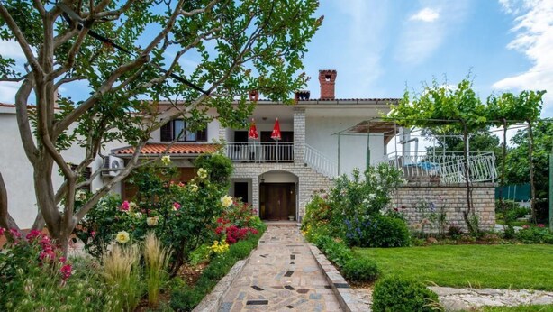 Una casa de dos pisos con un camino de piedra que lleva a la entrada, rodeada de exuberantes jardines y un árbol.