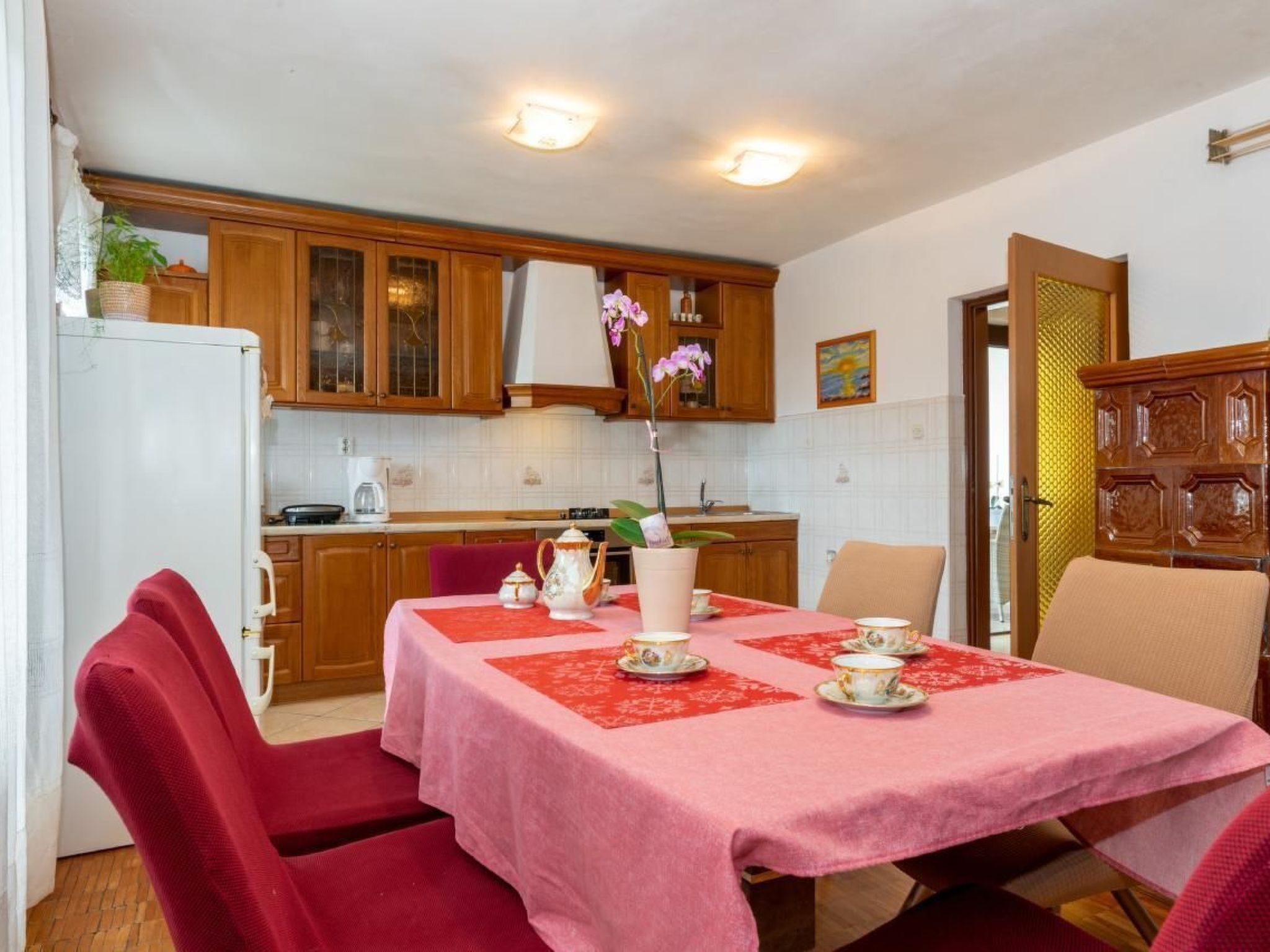 Un coin repas avec une table dressée pour quatre personnes, des meubles de rangement en bois, un réfrigérateur blanc et, au fond, une cuisine.
