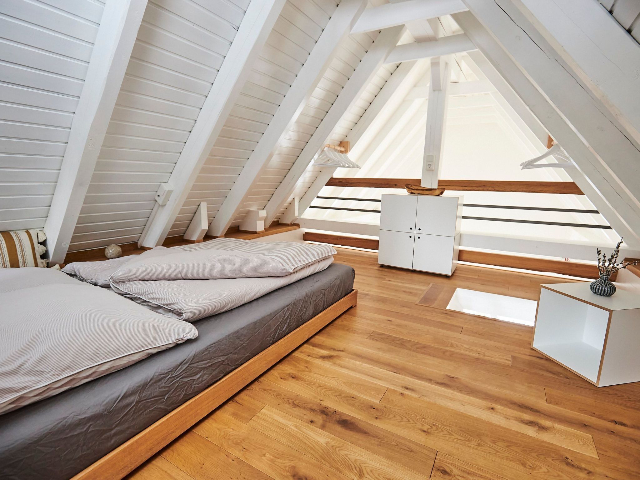 A loft bedroom with a wooden floor, a bed with pillows, a white wardrobe, and a small table with a vase.