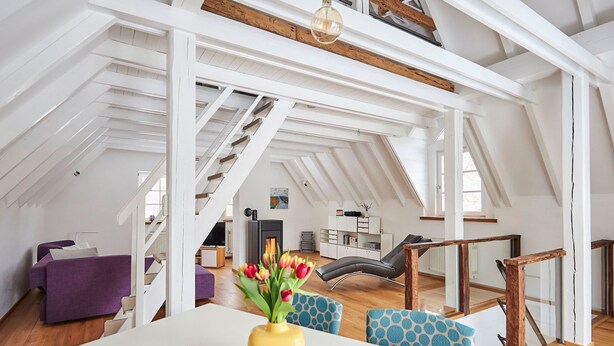 Ein großzügiges Loft mit hohen Decken, freiliegenden Holzbalken, einem zentralen Kamin und einem Essbereich mit Tisch und Stühlen.