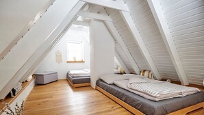 Ein Schlafzimmer im Loft-Stil mit schrägem Dach, Holzboden, einem Bett mit grauer Bettwäsche, einer kleinen Sitzecke und einem Fenster, das natürliches Licht hereinlässt.