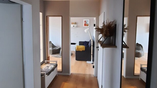 Um corredor com piso de madeira, paredes brancas e um espelho grande à direita. Há uma porta à esquerda e um quarto com uma cama e uma planta ao fundo.