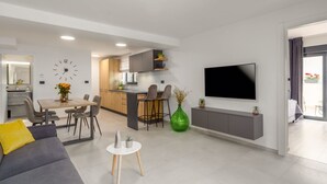 Un saló modern amb un sofà gris, una zona de menjador amb taula i cadires, una cuina amb un armari gris i un televisor penjat a la paret.