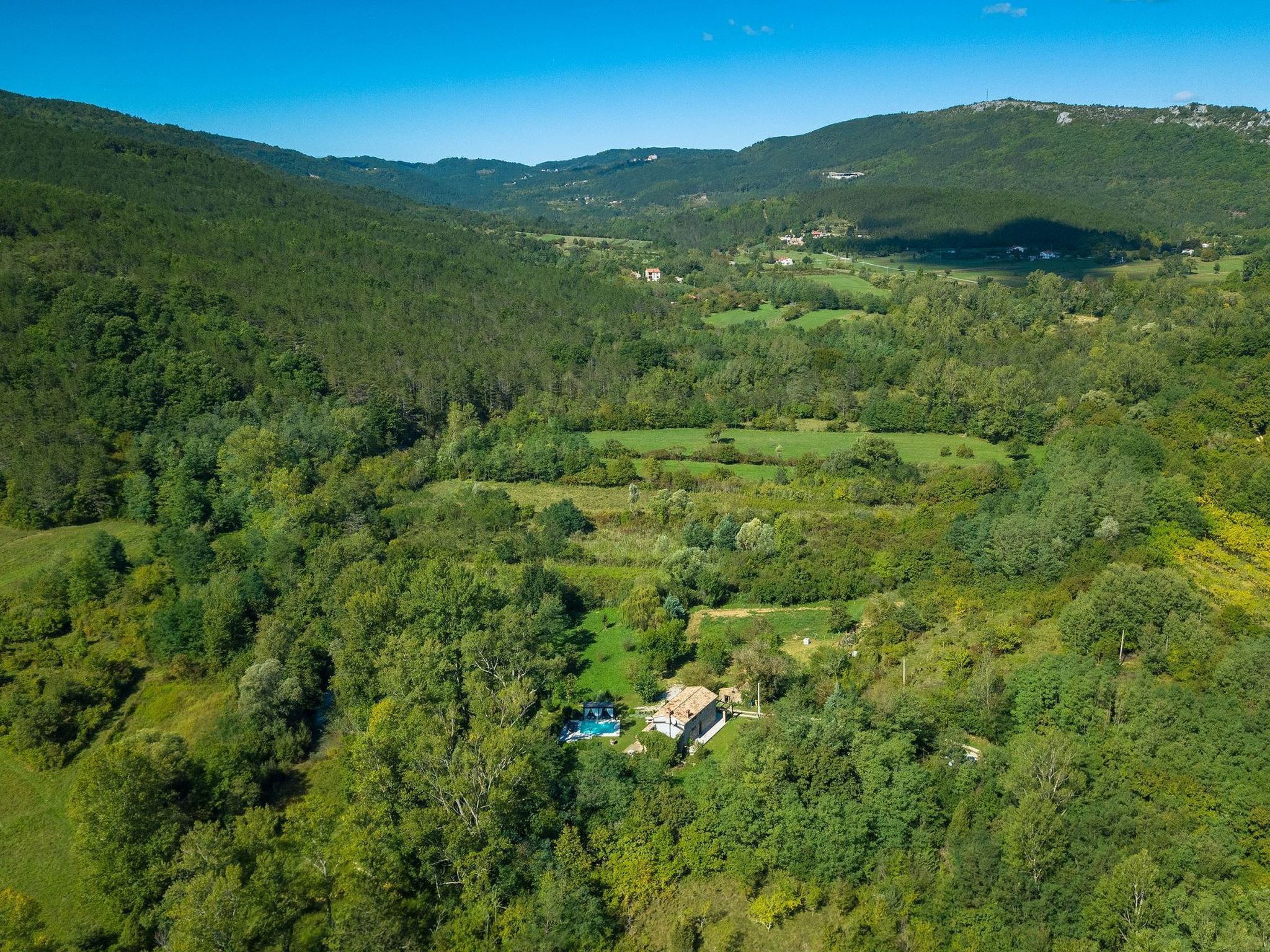 Vista aérea de un paisaje rural con una casa rodeada de bosque denso y colinas ondulantes.