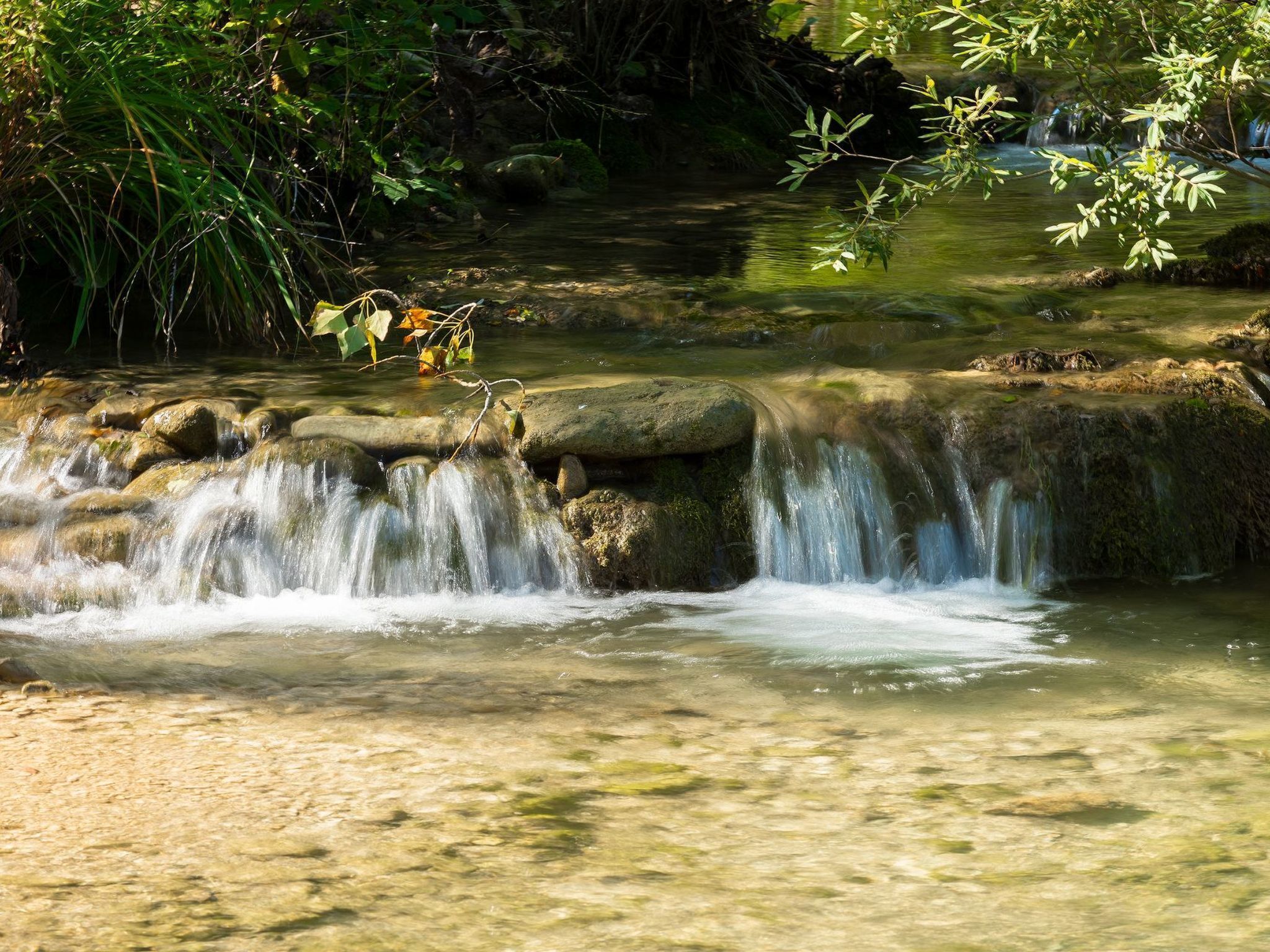 Ein natürlich entstandener Wasserlauf mit klarem Wasser, das über Steine fließt, umgeben von viel Grün.