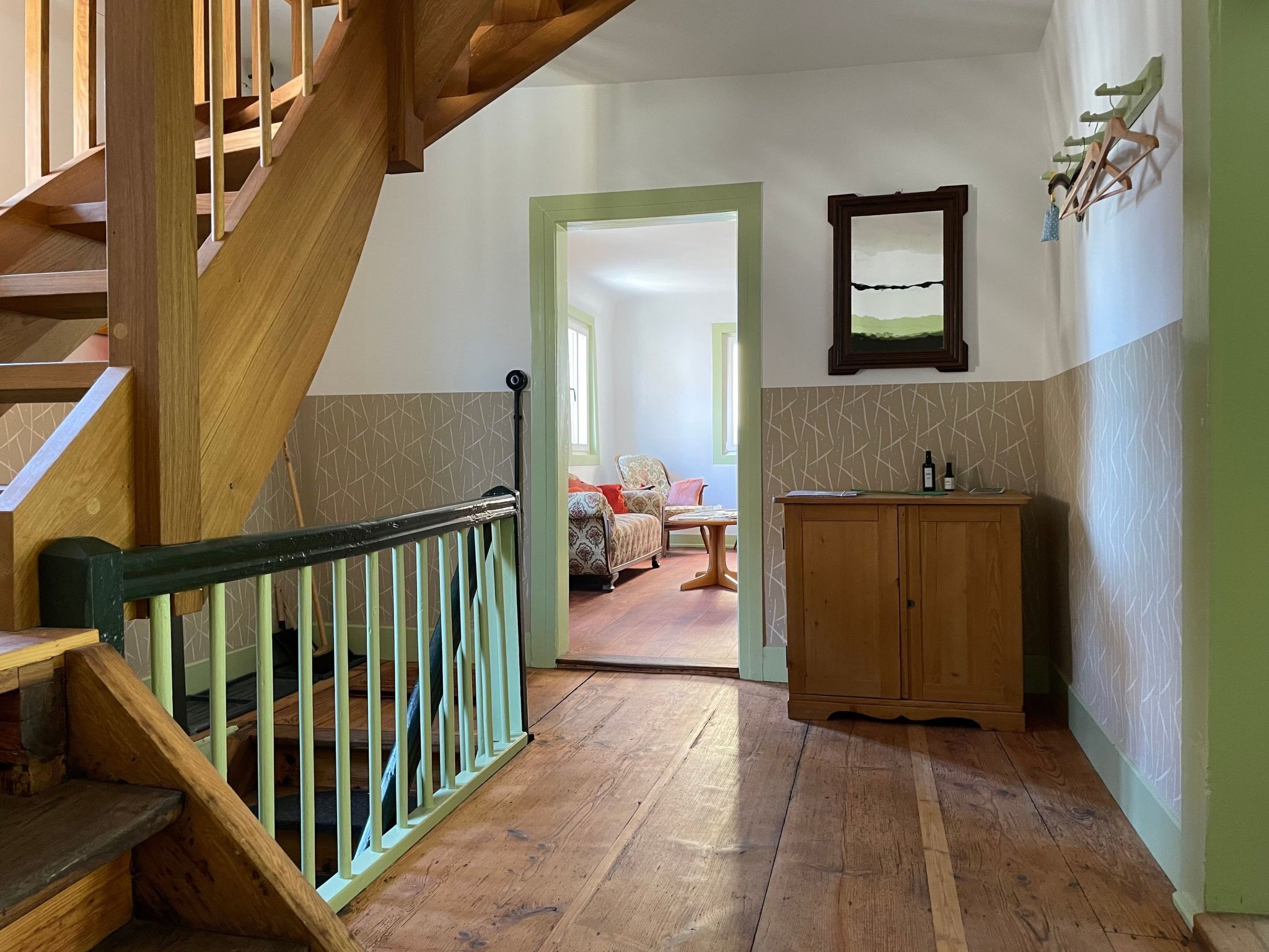 Eine Holztreppe mit grüner Handlauf, die zu einem Raum führt, der mit gemustertem Wandpapier, einem Holzschrank und einem gerahmten Bild dekoriert ist.