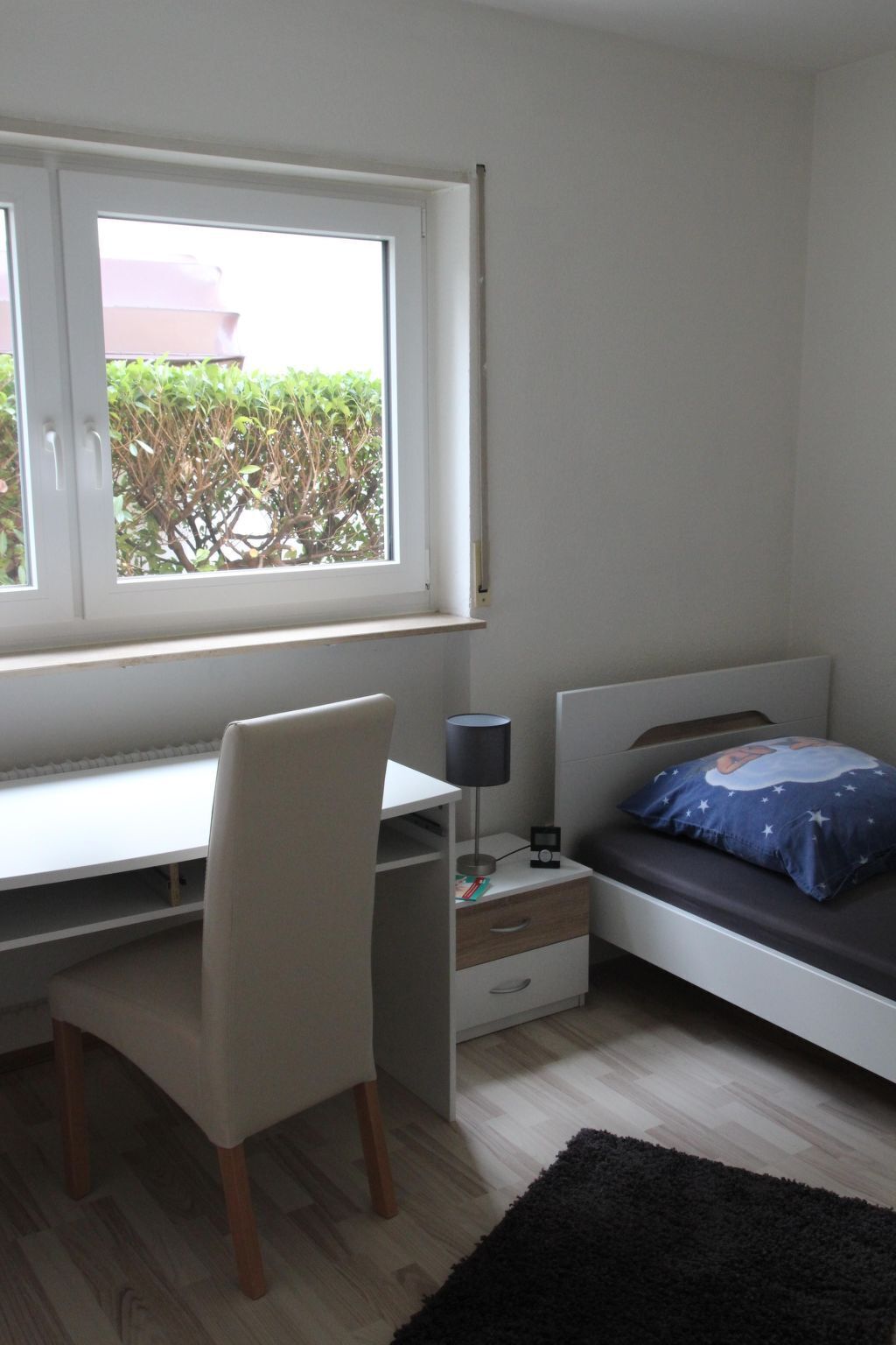 Un dormitorio con escritorio, silla, cama, mesita de noche, lámpara y vistas al exterior con vegetación.