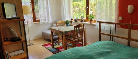 A cozy room with a bed, a desk, and a chair. There is a window with curtains, a plant, and a small table with decorative items.