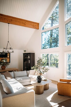 A spacious living room with a high ceiling, large windows, a wooden ceiling beam, a modern chandelier, and a comfortable seating area. - Prow & Pines A-Frame: Hot Tub • EV • Lake Rights (Lake Arrowhead)