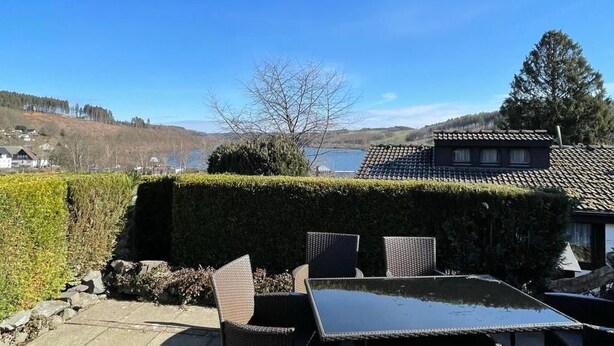 Eine Terrasse im Freien mit Korbmöbeln und Blick auf einen See und Hügel.