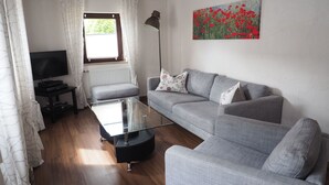 A living room with a grey sofa, a glass coffee table, a black TV stand, and a floor lamp. There is a window with curtains and a painting of red flowers on the wall. - Hochwertige Ferienwohnung mit Terrasse und Garten by Interhome (Zülpich)