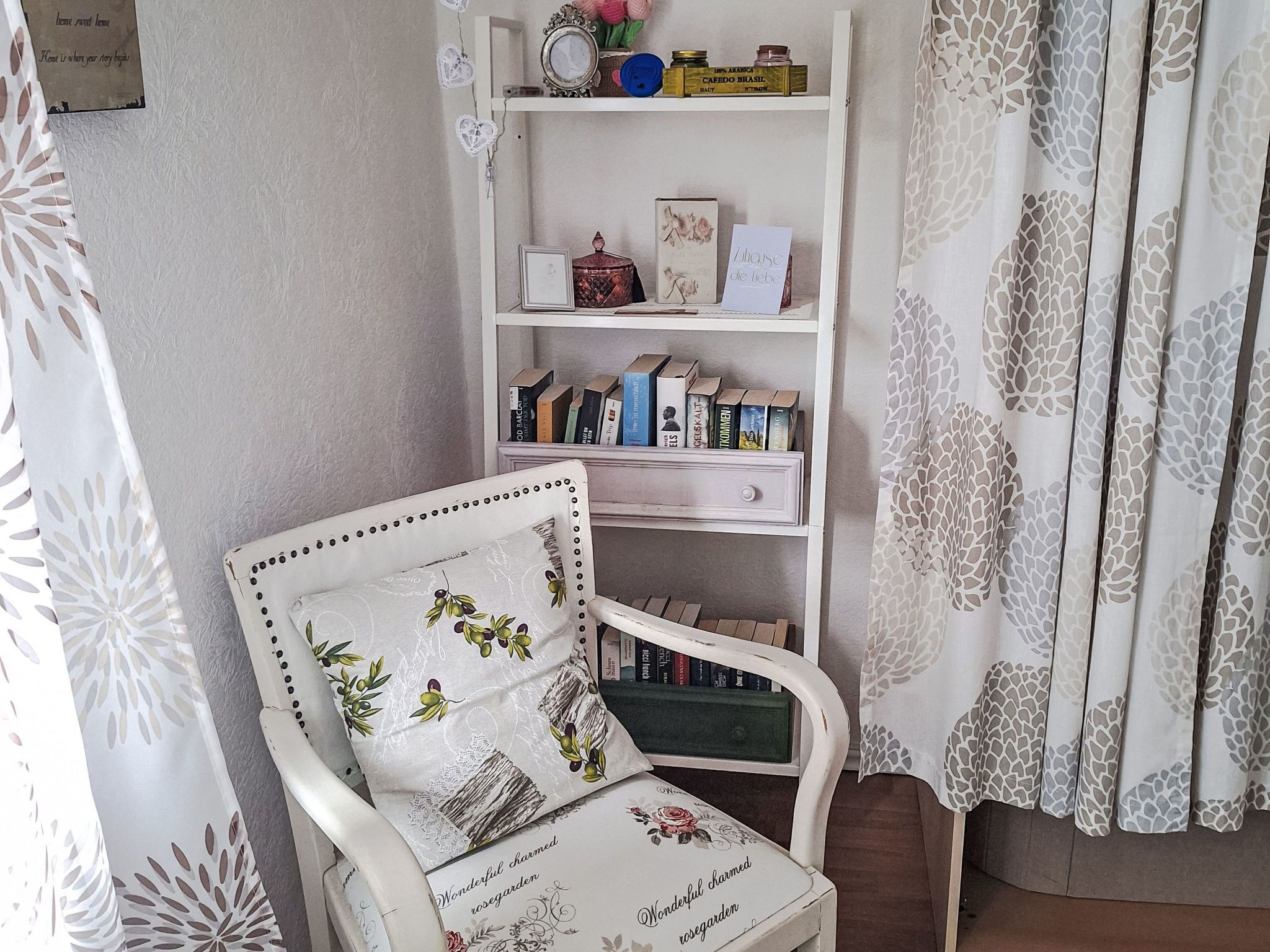 A white armchair with floral upholstery, a bookshelf with books and decorative items, and a patterned curtain.