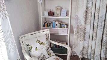 A white armchair with floral upholstery, a bookshelf with books and decorative items, and a patterned curtain.