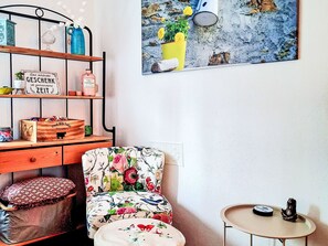 A room with a wooden desk, a chair with floral upholstery, a small round table, and a wall-mounted shelf with various items. - Ferienwohnung Sander by Interhome (Kirchen)