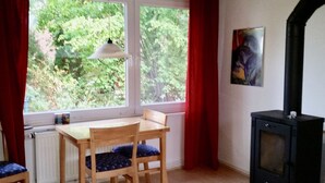 Una habitación acogedora con una mesa y sillas de madera, un horno de leña negro, un radiador y una ventana con cortinas rojas.