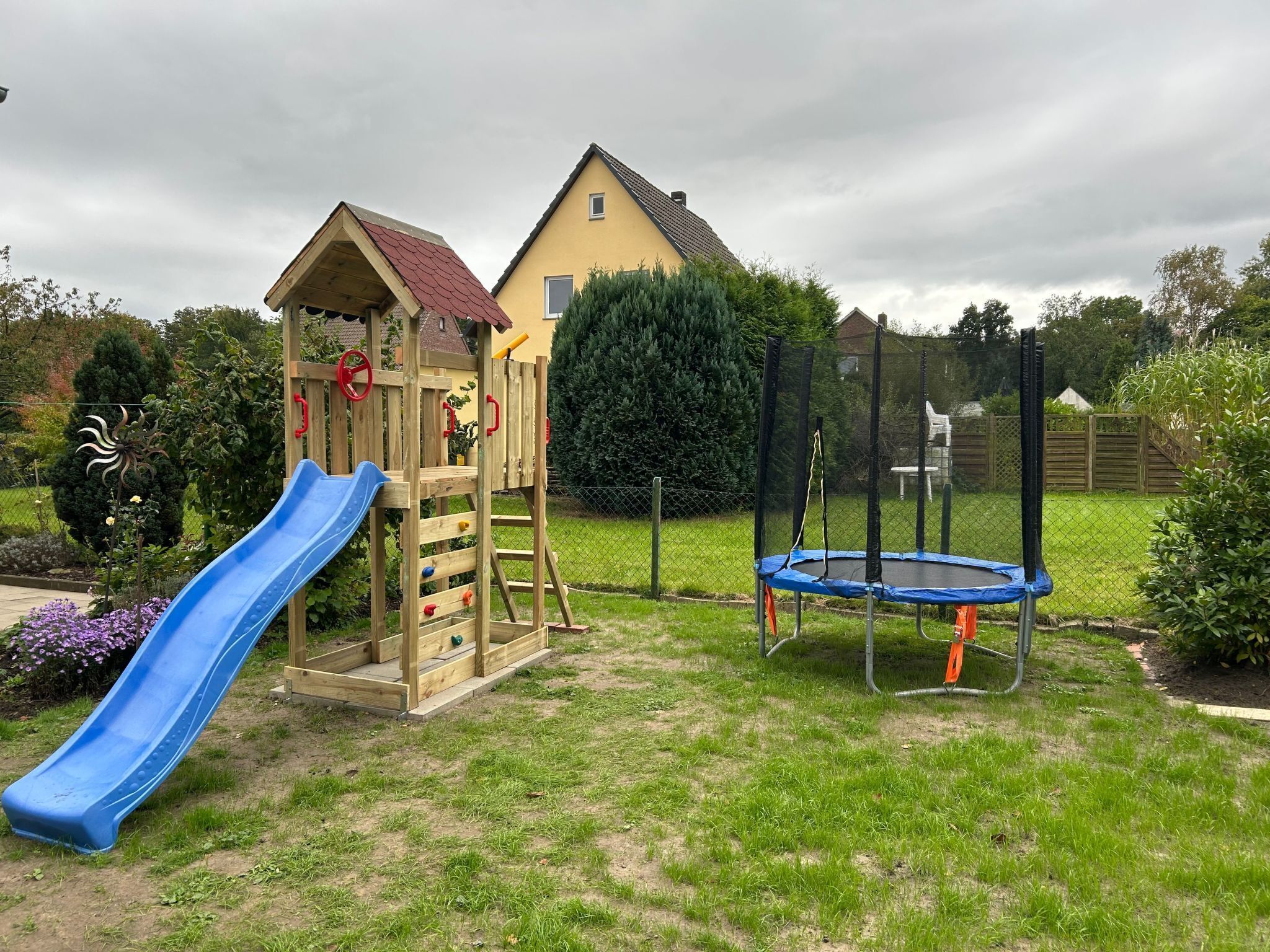 A playground with a slide and a trampoline, surrounded by a fence and greenery.