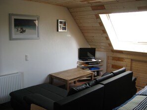 Una habitación acogedora con techo inclinado, una mesa de madera, un sofá negro y un televisor sobre una estantería.