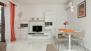 Ein modernes Wohnzimmer mit Fernseher, weißen Möbeln, einem Esstisch mit Stühlen und einem Bild an der Wand.